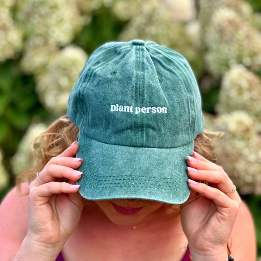 Woman with curly hair and manicured nails wearing a washed green denim baseball cap that says plant person in white font