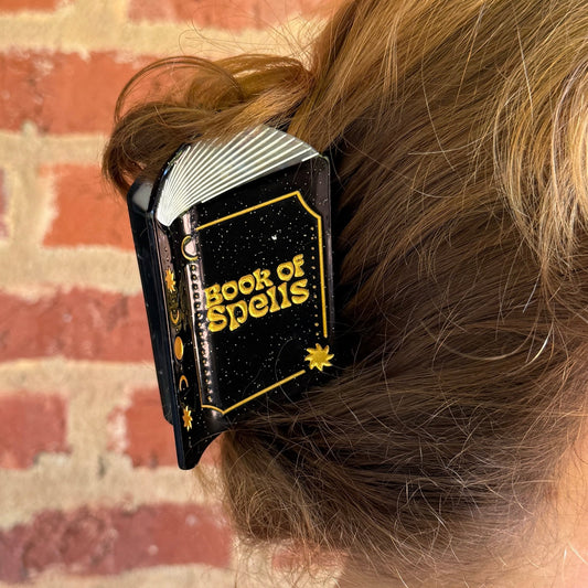 A close-up of a person's light brown hair held in a black hair clip with gold lettering that reads 'BOOKS OF SPELLS'. The background shows a brick wall.