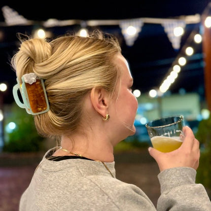 Woman holding a glass of beer with a mug-shaped hair clip in her hair, standing outdoors at night.