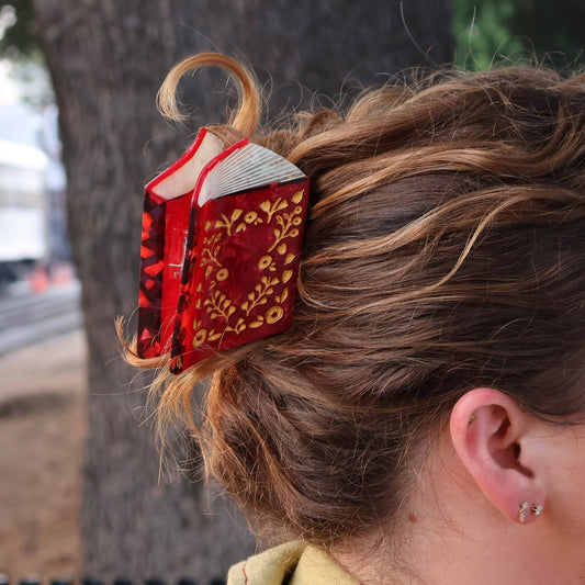 A red and gold book-shaped hair claw clip holding up a woman's hair outdoors, with a tree trunk blurred in the background.