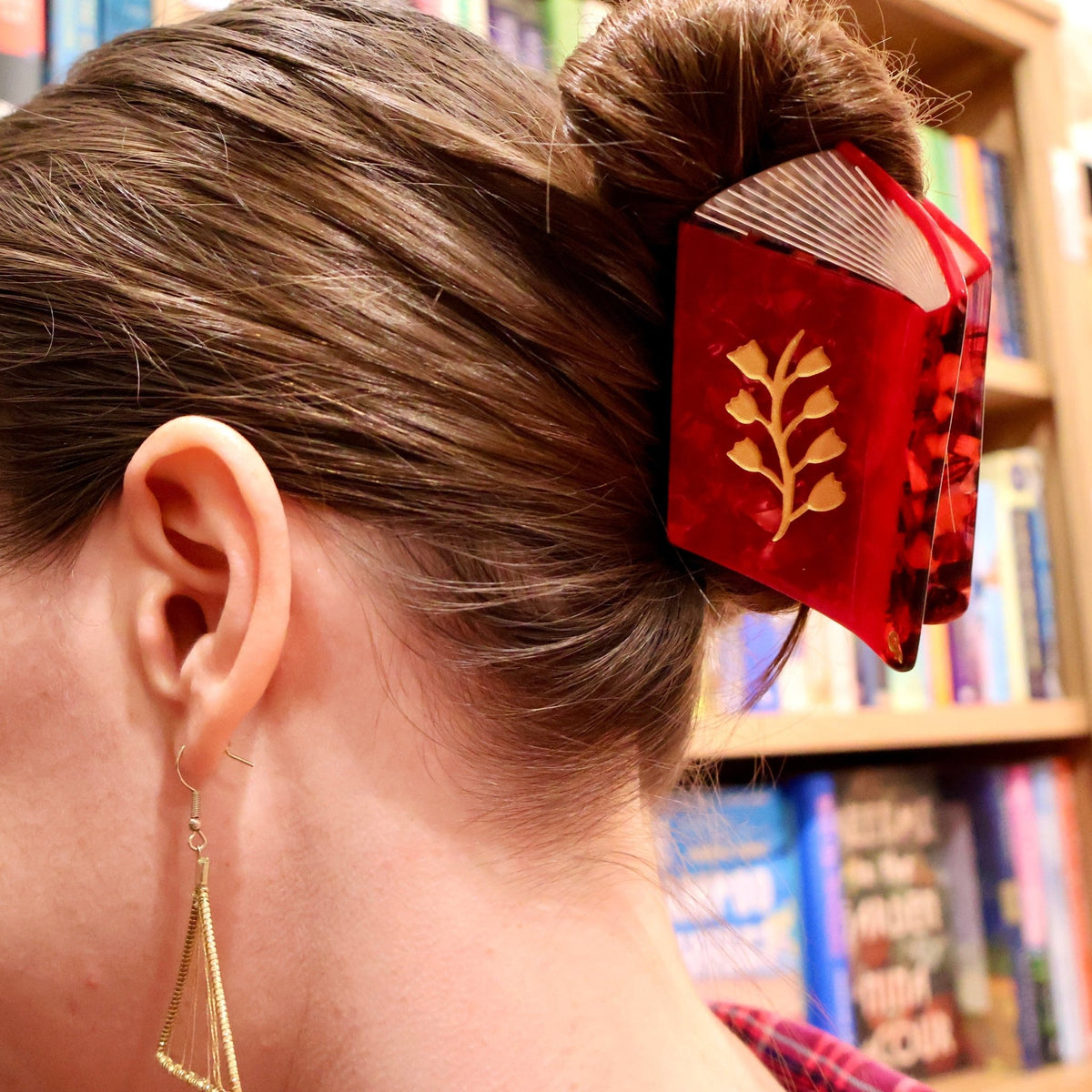 A close-up side view of a woman with her hair in a bun, wearing a red hair accessory shaped like a small book with gold leaf detailing, set against a blurred background of bookshelves.
