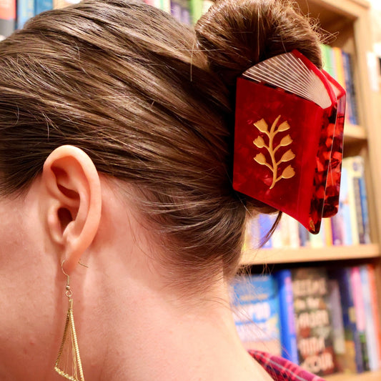 A close-up side view of a woman with her hair in a bun, wearing a red hair accessory shaped like a small book with gold leaf detailing, set against a blurred background of bookshelves.