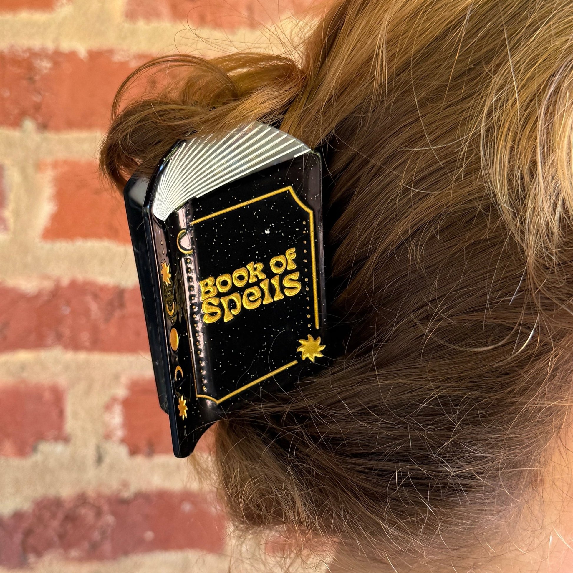 A close-up of a person's light brown hair held in a black hair clip with gold lettering that reads 'BOOKS OF SPELLS'. The background shows a brick wall.
