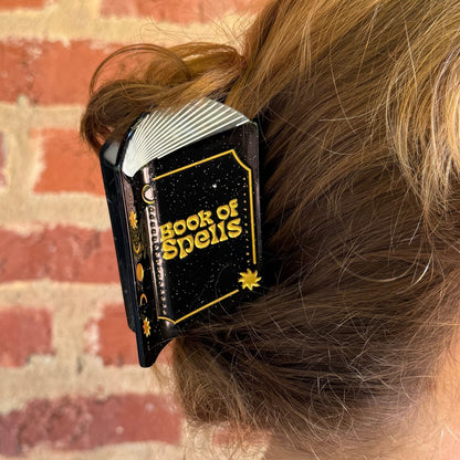 A close-up of a person's light brown hair held in a black hair clip with gold lettering that reads 'BOOKS OF SPELLS'. The background shows a brick wall.