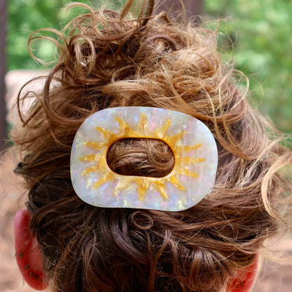 A close-up lifestyle shot of a person's messy brown hair secured with a white and yellow sun-shaped hair claw clip outdoors.
