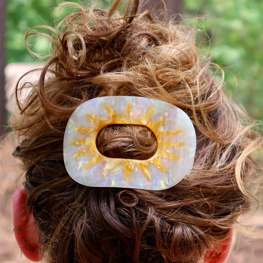 A close-up lifestyle shot of a person's messy brown hair secured with a white and yellow sun-shaped hair claw clip outdoors.