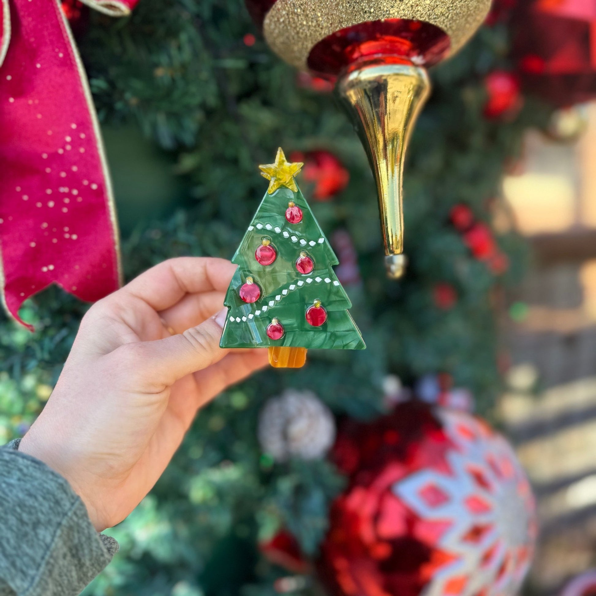 A hand holds a small green Christmas tree ornament with red ornaments and a gold star, in front of a decorated Christmas tree with red and gold ornaments.