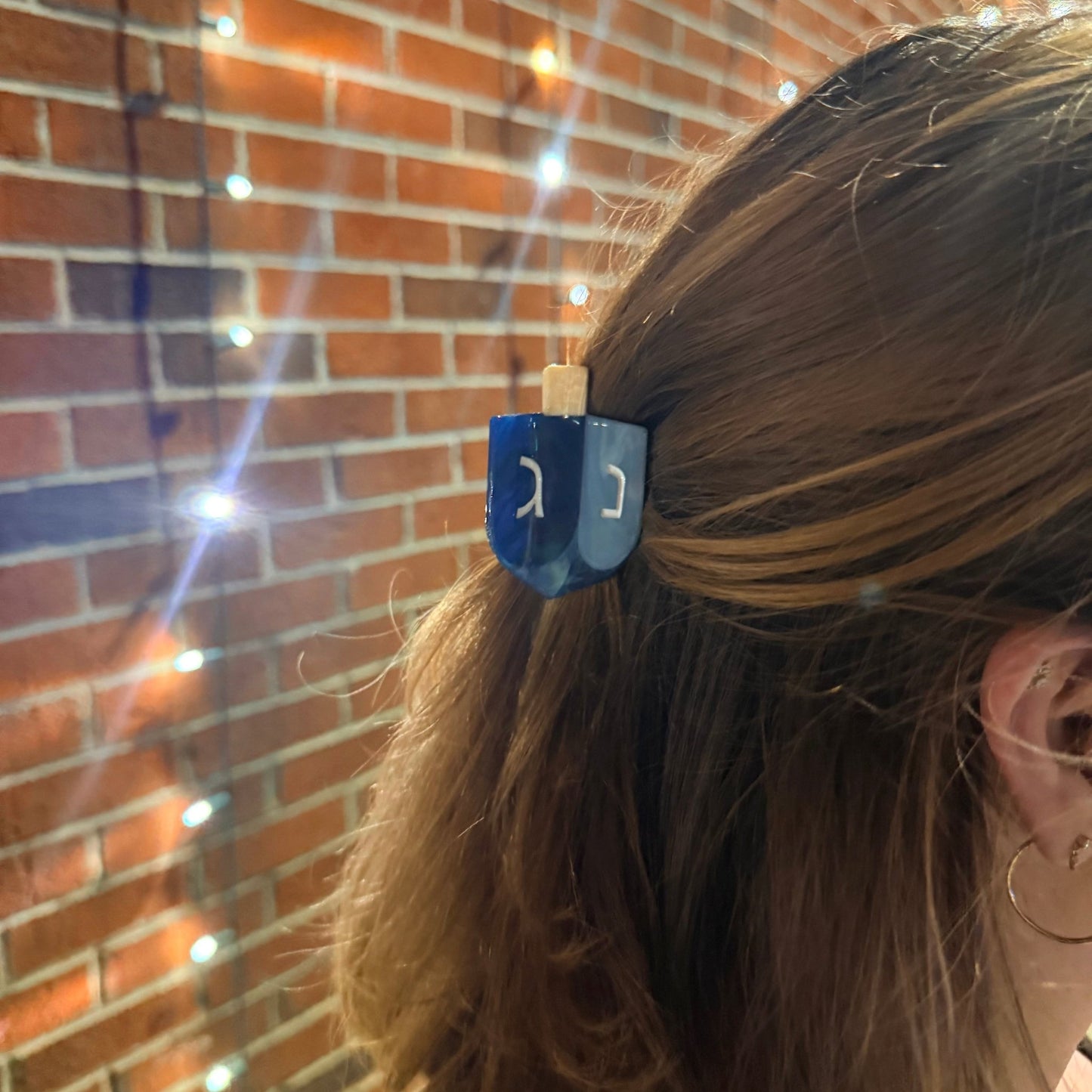 A close-up lifestyle shot of a woman's brown hair secured with a blue dreidel hair clip against a warm brick wall with glowing fairy lights.