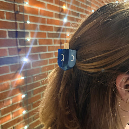 A close-up lifestyle shot of a woman's brown hair secured with a blue dreidel hair clip against a warm brick wall with glowing fairy lights.