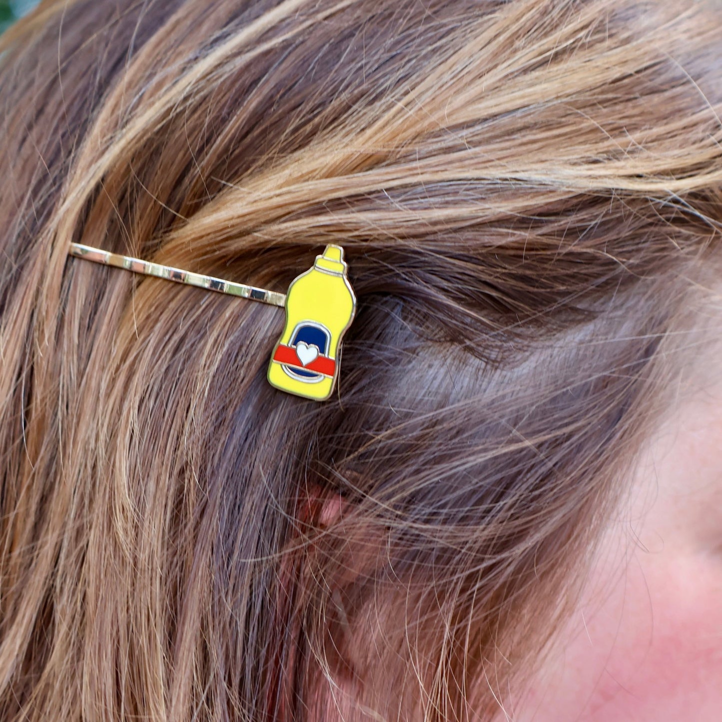 A close-up lifestyle shot of a person's brown hair wearing a gold bobby pin hair clip shaped like a yellow mustard bottle with a heart label.