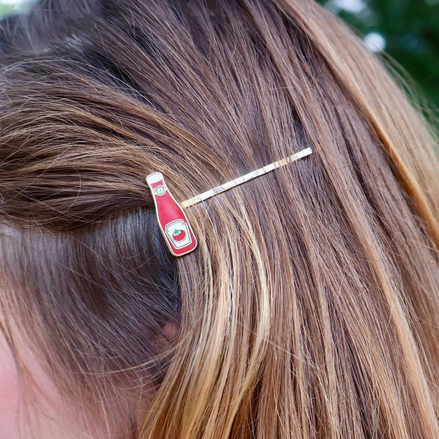 A close-up of a person's brown hair secured with a small hair clip shaped like a red bottle with a gold-tone pin.