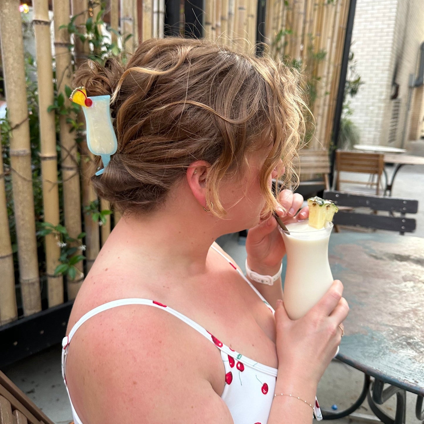 A woman in a white cherry-print dress and a pina colada shaped hair claw clip holding a tropical cocktail glass with a straw, sitting at an outdoor patio table.