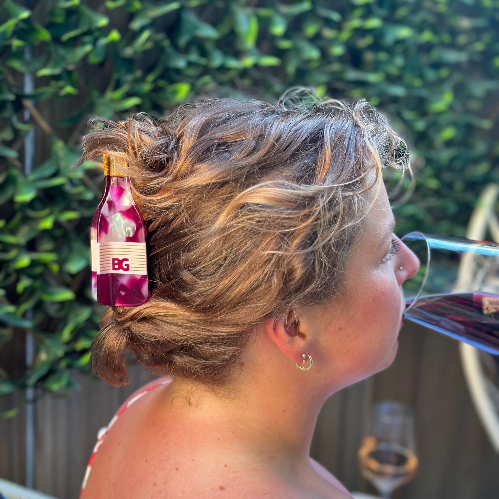 A woman in profile outdoors with her hair in a messy bun, wearing a small burgundy wine bottle hair clip with a pink and white floral pattern and a gold cap. She is drinking from a glass of red wine, with a blurred green hedge in the background.