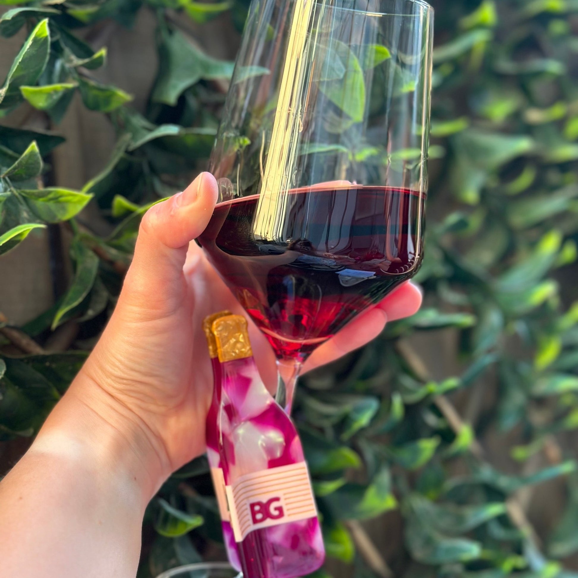 A hand holding a wine glass filled with red wine, with a small bottle of BG red wine in the foreground and a green leafy hedge in the background.