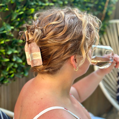 A woman outdoors wearing a large, light pink wine bottle claw clip hair accessory that holds her messy blonde-up styled hair in place. She is holding a glass of rosé wine, with a wooden fence and green foliage in the background.